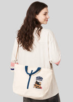 Via Artists Ikaku Goron 2-way cotton canvas tote bag with adjustable strap and embroidered graphic, worn over shoulder.